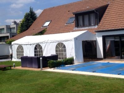 Un grand chapiteau blanc installé dans le jardin d'une maison résidentielle. Le chapiteau est placé à côté d'une piscine rectangulaire, avec une couverture de piscine bleue partiellement visible. La maison a un toit en tuiles rouges et des fenêtres à cadre sombre. Le jardin est bien entretenu avec une pelouse verte et des meubles de jardin, créant un cadre agréable pour un événement en plein air.