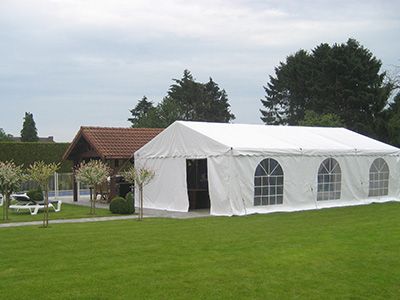 Un grand chapiteau blanc installé sur une pelouse verte bien entretenue. Le chapiteau a des fenêtres en forme d'arcs sur les côtés, laissant entrer la lumière naturelle. À proximité, il y a une maison avec un toit en tuiles rouges et des arbres bien taillés. Le chapiteau semble prêt pour un événement en plein air.